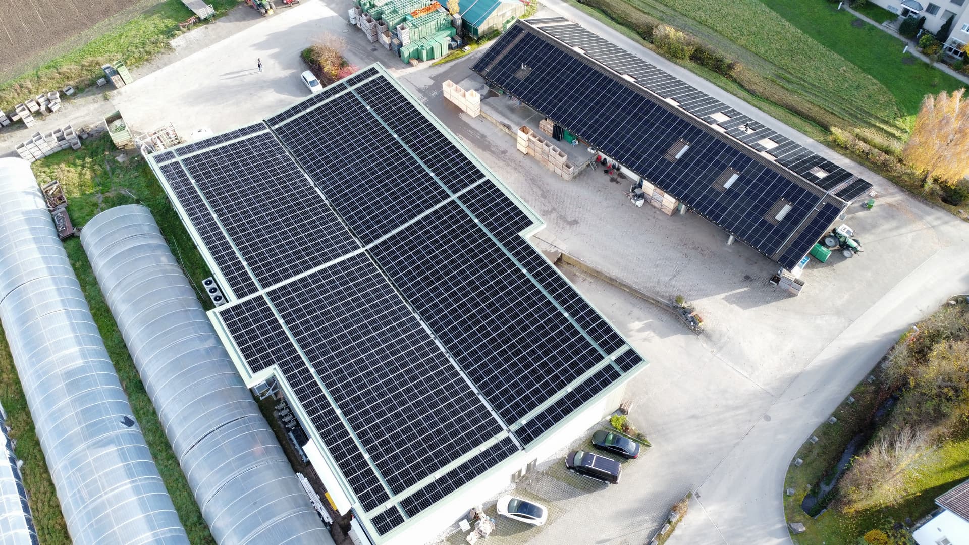 Luftaufnahme von Photovoltaikanlagen auf zwei Betriebsgebäuden neben Gewächshäusern